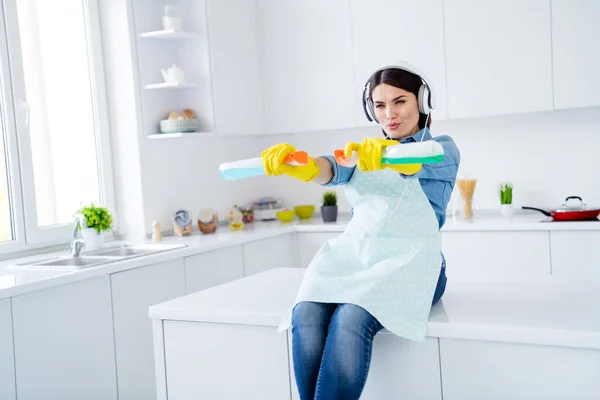 Portrait of positive funky funny playful woman sit table hold sprayer bottle spray disinfect kitchen corona virus listen headset music song sound in house indoors — Stock Photo, Image