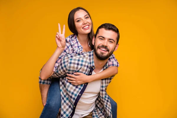 Foto de linda senhora bonito cara casal se divertindo segurando as mãos piggyback mostrando símbolo v-sinal alegre bom humor desgaste casual xadrez camisas jeans isolado cor amarela fundo — Fotografia de Stock