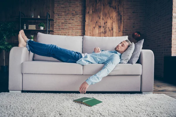 Full body profile photo of domestic handsome guy stay home quarantine time fall asleep reader book on floor lying barefoot cozy sofa daydream modern interior living room indoors