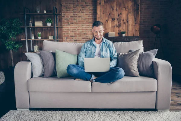 Foto de chico guapo y hogareño relajante sentado cómodo sofá piernas cruzadas portátil de navegación freelancer trabajo remoto quedarse en casa buen humor cuarentena tiempo sala de estar en el interior —  Fotos de Stock