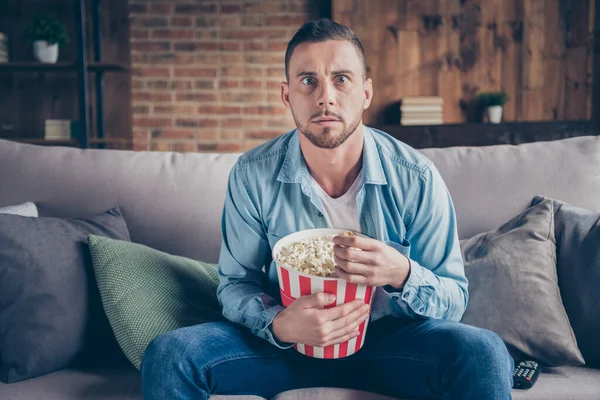 Foto de chico guapo doméstico relajante estancia en casa cuarentena tiempo ver televisión película de terror comer palomitas de maíz aterrorizado sentarse acogedor sofá moderno interior sala de estar interior — Foto de Stock