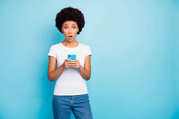 Foto de estupor bela pele escura ondulada senhora segurando telefone app mãos boca aberta ler cama notícias desgaste casual branco t-shirt jeans isolado azul cor fundo — Fotografia de Stock