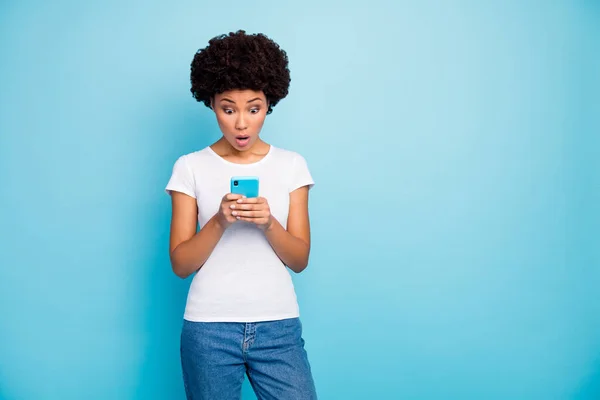 Foto de sem palavras bela pele escura ondulada senhora segurando as mãos do telefone boca aberta ler cama notícias desgaste casual branco t-shirt jeans isolado azul cor fundo — Fotografia de Stock