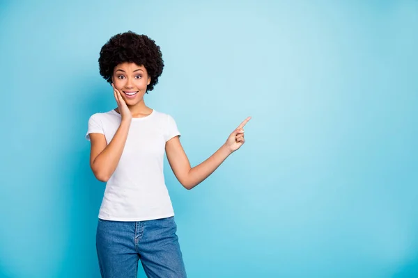 Foto van grappige vrij donkere huid krullende dame sales manager aangeeft vingers lege ruimte voorstellen lage prijzen dragen casual witte t-shirt jeans geïsoleerde blauwe kleur achtergrond — Stockfoto