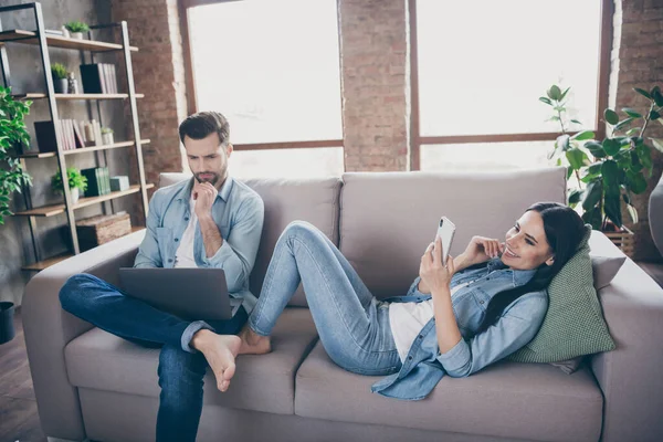 Foto de dos personas cónyuges hombre sentarse sofá trabajo portátil remoto analizar documento mujer mentira uso teléfono inteligente chatear mensajes de texto escribiendo en casa en el interior — Foto de Stock