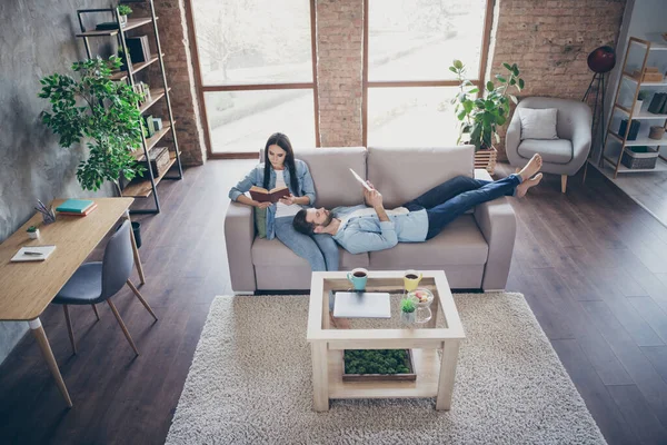 Сверху над высоким углом зрения полный размер фото сфокусированной супружеской пары женщина сидеть читать бумажную книгу мужчина лежит диван смотреть табличку в доме внутри — стоковое фото