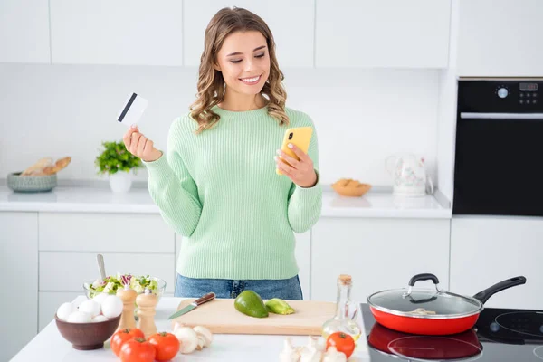 Portrait of her she nice attractive lovely cheerful girl cooking fresh tasty yummy meal holding in hands ell bank card ordering vegs farm organic products in modern light white kitchen house