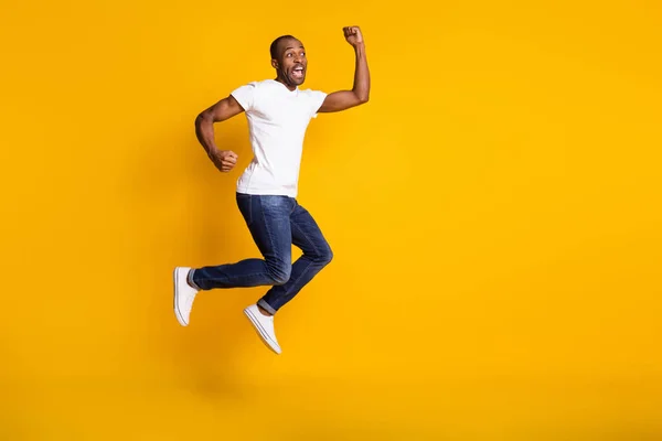 Full storlek foto av attraktiv mörk hud kille hoppar högt upp glädje bäst vinna sportspel bookmaker bära casual vit t-shirt jeans skor isolerad gul färg bakgrund — Stockfoto