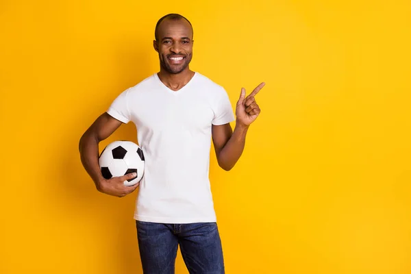 Фото привлекательной темной кожи парень держит кожаный футбольный мяч хорошее настроение наркоман вентилятор прямой палец пустое пространство носить повседневные джинсы белые футболки изолированные желтый яркий цвет фона — стоковое фото