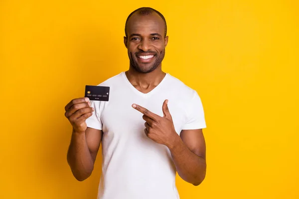 Retrato de positivo afro-americano cara segurar cartão de crédito sugerem fácil bancário lucro ponto de pagamento dedo indicador desgaste branco t-shirt isolado sobre brilhante brilho cor fundo — Fotografia de Stock