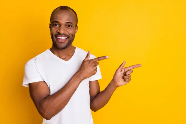 Foto di attraente pelle scura ragazzo tenere le braccia dirigendo dita lato spazio vuoto che mostra novità informazioni indossare casual bianco t-shirt isolato luminoso vivido colore giallo sfondo — Foto Stock