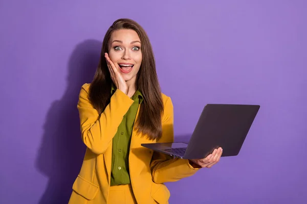 漂亮的女员工离家工作在家用笔记本电脑工作的照片拿到了很高的薪水不相信眼睛手臂贴在脸颊上穿着黄色西服孤立的紫色彩色背景 — 图库照片