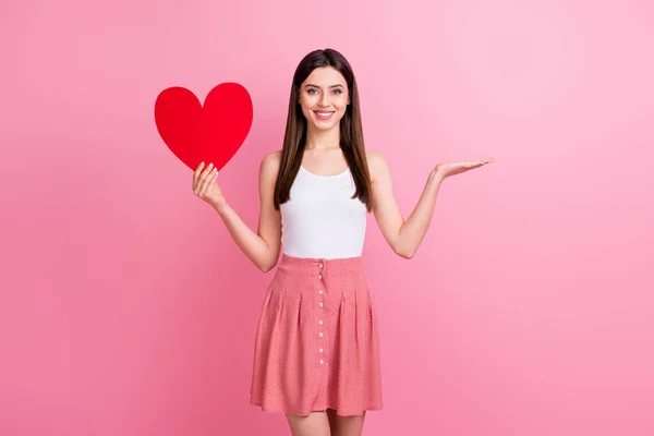 Foto de senhora bonita segurar grande papel vermelho coração mão mostrando no braço aberto novidade surpresa produto desgaste casual branco singlet curto pontilhado saia isolado pastel cor rosa fundo — Fotografia de Stock