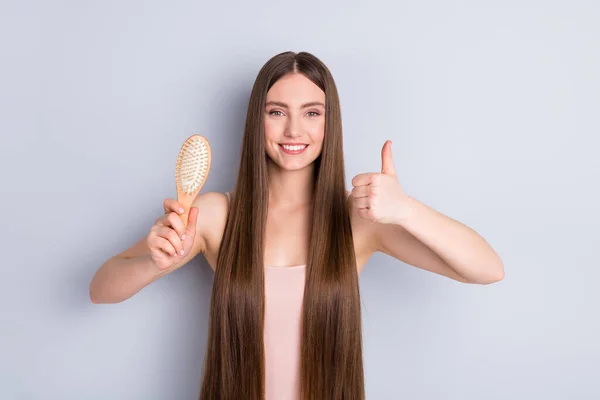 Photo of charming wygląd model lady demonstrujące idealne schludne długie fryzury pokazując ulubione drewniane grzebień podnieść palec kciuk do góry nosić singlet odizolowany szary kolor tła — Zdjęcie stockowe