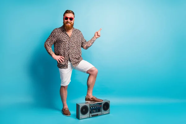 Full length photo of macho guy one leg stand on retro tape recorder direct finger empty space advice novelty wear shorts leopard shirt footwear sun specs isolated blue background — Stock Photo, Image