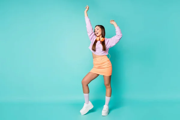 Foto de tamaño completo de linda dama milenaria escuchar los auriculares de la tecnología moderna danza alegre levantar las manos desgaste recortado suéter violeta falda naranja zapatillas aisladas color verde azulado fondo — Foto de Stock