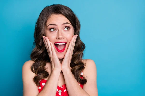 Closeup photo of pretty funny shocked wavy lady arms on cheeks open mouth excited overjoyed expression look side empty space wear red dotted dress singlet isolated blue color background