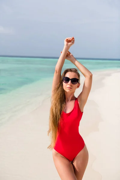魅力的な女性の垂直写真ロングヘア堂日光浴スタンド砂フィット図海辺のエキゾチックなリゾート残りの部分は腕を上げる太陽の仕様を着用赤いボディスーツ外暖かい太陽光線海のビーチを楽しむ — ストック写真