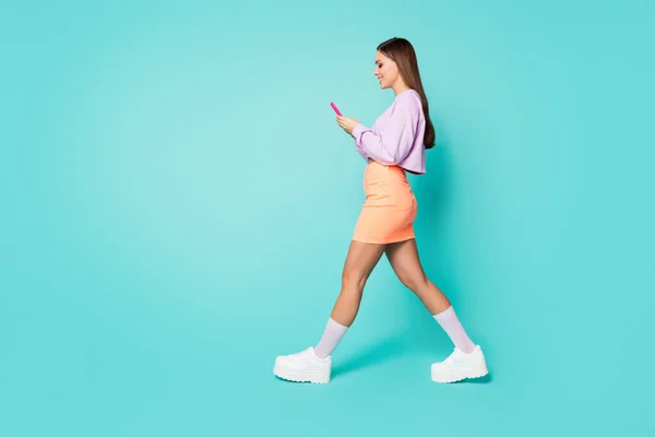 Full body profile photo of cool lady youth outfit hold telephone hands walk street write message wear cropped pullover orange skirt shoes long socks isolated teal color background