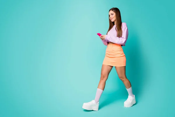 Full längd profil foto av funky cool dam ungdom outfit hålla telefon promenad gata slitage trend lila beskärda tröja naken mage orange kjol skor strumpor isolerad teal färg bakgrund — Stockfoto