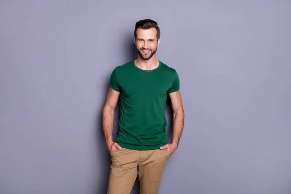 Photo of attractive guy business man perfect schludny fryzura szczecina wygląd uśmiechnięty belka dobry nastrój ręce kieszenie nosić casual zielony t-shirt spodnie odizolowany szary kolor tła — Zdjęcie stockowe