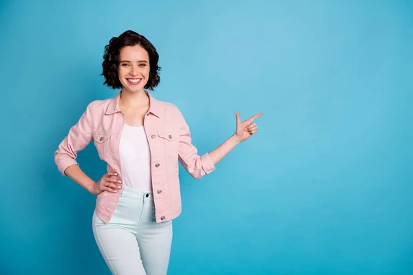 Photo of attractive lady short black hairdo beaming smiling good mood direct finger side empty space show pandemia rules wear casual pink denim jacket pants isolated blue color background — Zdjęcie stockowe