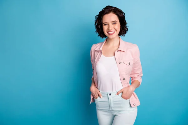 Foto de senhora bonita atraente curto penteado preto com raios de dentes sorrindo braços de bom humor em bolsos rua olhar desgaste casual rosa calça jeans casaco isolado azul cor de fundo — Fotografia de Stock