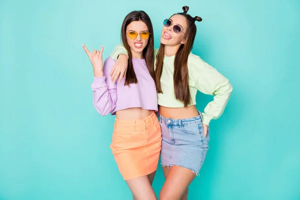 Photo of two funny crazy behavior ladies show horns hands stick tongue out mouth wear sun specs green violet pullovers naked belly mini skirt isolated pastel teal color background — Stock Photo, Image