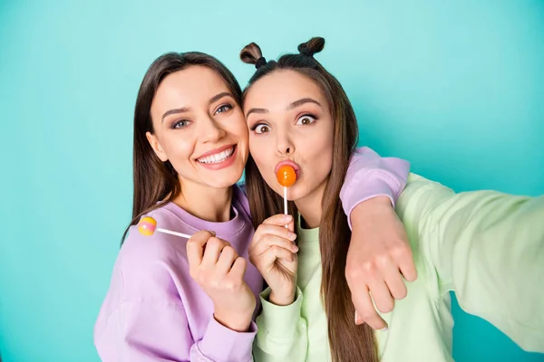 Foto van twee grappige dames houden lolly chupa chups handen kinderachtig lekker snoep maken selfies dragen groen violet pullovers geïsoleerde pastel teal kleur achtergrond — Stockfoto