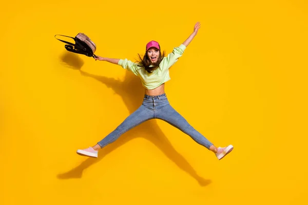 Foto corpo pieno di funky energico signora studente universitario tenere lo zaino salto in alto esultante usura verde felpa tagliata jeans scarpe cap isolato vivido colore giallo brillante sfondo — Foto Stock