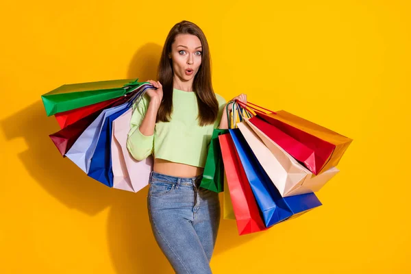 Foto de la encantadora señora sorprendida buen humor calle mirada celebrar muchas bolsas de compras venta de la boca abierta desgaste verde recortado sudadera jeans aislados vivos de color amarillo brillante fondo —  Fotos de Stock