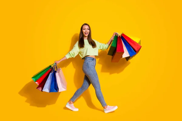 Full size foto profilo di bella signora shopaholic buon umore camminare negozio centro commerciale tenere molte borse indossare verde felpa ritagliata jeans scarpe isolato vivido colore giallo brillante sfondo — Foto Stock