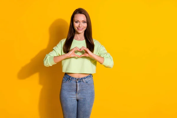 Foto de atractiva señora bonita de buen humor sostener los brazos haciendo figura del corazón forma llena de alegría romance desgaste verde recortado sudadera jeans aislados vivos de color amarillo brillante fondo —  Fotos de Stock