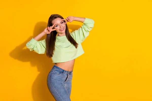 Foto di attraente piuttosto divertente signora buon umore braccia che mostrano v-segno simboli vicino agli occhi flirty usura felpa ritagliata verde maglione jeans isolato vivido colore giallo brillante sfondo — Foto Stock