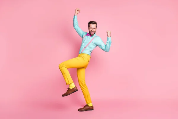 Teljes hosszúságú fotó őrült boldog jóképű srác trend ruhák emelje öklöt fel hurrá viselni rózsaszín ing nadrágtartó csokornyakkendő sárga nadrág cipő elszigetelt pasztell rózsaszín szín háttér — Stock Fotó
