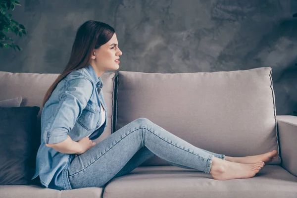 Полная длина профиля боковой фото разочарованной больной девушки сидеть диван прикоснуться к животу у нее проблемы с пищеварением менструации пищевого отравления одежда джинсы в доме в помещении — стоковое фото