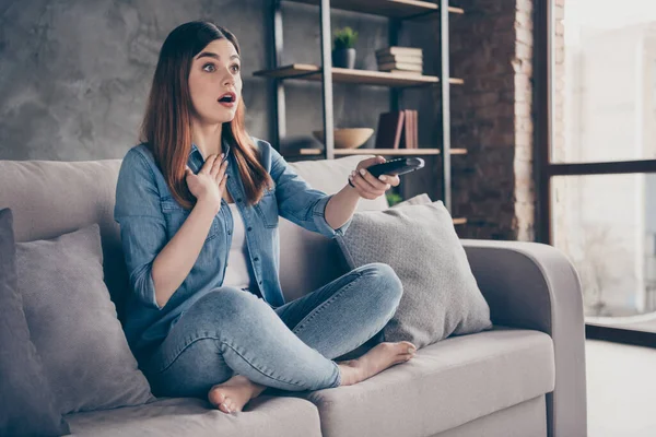 Paura ragazza sit divan gambe incrociate tenere telecomando guardare la tv stupito incredibile horror serie finale toccare mano petto in camera appartamento al chiuso — Foto Stock