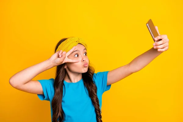 Foto de divertida señorita mantener el teléfono cuarentena tiempo quedarse en casa hablar skype compañeros de clase mostrar v-signo cerca de ojo enviar besos usar casual azul camiseta diadema aislado color amarillo fondo — Foto de Stock