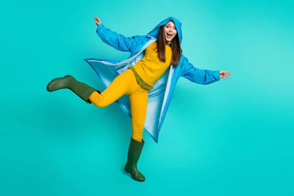 Foto de corpo inteiro de atraente engraçado senhora alegre humor chuvoso tempo rua passeio capuz na cabeça muitas poças levantar perna desgaste capa de chuva calças chiclete botas isolado cor teal fundo — Fotografia de Stock