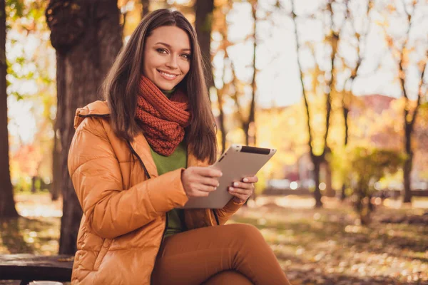 웃고 있는 매력적 인 아가씨가 벤치 공원에 앉아 있는 사진기다리고 있는 남자 친구가 쓰고 있는 타블렛 사진들이 공개 된 가을 사진들은 스카프그린 터틀넥 오렌지 바람막이 바지를 착용하고 있다. — 스톡 사진