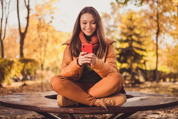 아름다운 여인이 의자에 앉아 미소를 짓고 있는 사진, 전화가 걸려 있고, 가을 사진 친구의 소셜 네트워크 도시 공원은 스카프그린 터틀넥 주황색 바람막이 바지를 밖에서 입고 있다. — 스톡 사진