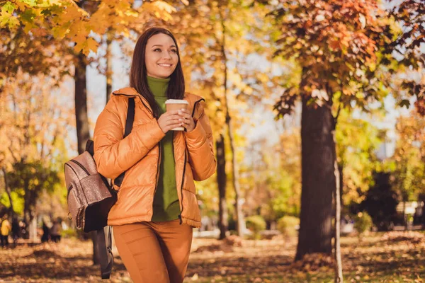 손으로 웃고 있는 매력적 인 아가씨가 웃으며 커피를 마시며 아침 가을 공원에 떨어지는 것을 보고 있는 이 사진은 반짝이는 배낭처럼 반짝이는 녹색 터틀넥 오렌지 바람막이 바지를 밖에 입고 있다 — 스톡 사진