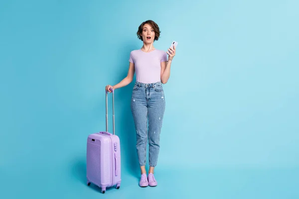 Full body photo of astonished girl tourist hold luggage use cellphone impressed quarantine border taxi hotel close wear lilac violet denim jeans isolated blue color background — Stock Photo, Image