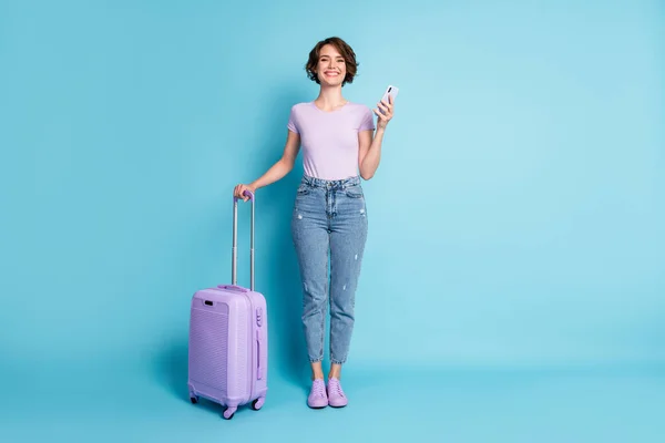 Foto de tamaño completo de chica alegre positivo equipaje de bodega turística uso smartphone reserva taxi hotel desgaste lila violeta camiseta vaqueros aislados color azul fondo —  Fotos de Stock