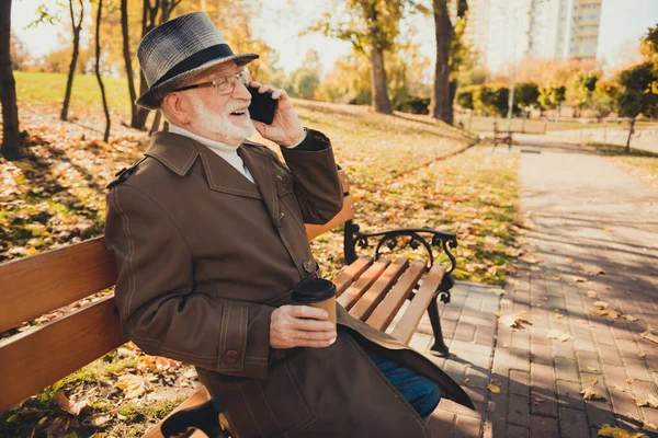 Perfil lado foto de gris blanco pelo viejo hombre sentarse banco llamada amigo teléfono inteligente celebrar bebida para llevar taza de café disfrutar de otoño naturaleza descanso relajarse parque usar chaqueta sombrero — Foto de Stock