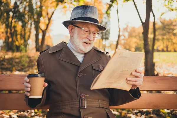 Photo de vieux pensionné excité repos repos se détendre automne parc assis banc lire journal boire emporter café boisson tasse porter veste chapeau casque manteau — Photo