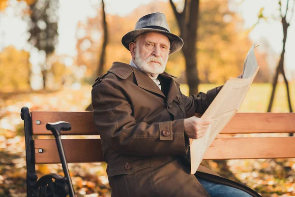 Photo of serious old man grumpy pensioner dont like kids play autumn town park sit bench read look newspaper wear cap long jacket outerwear