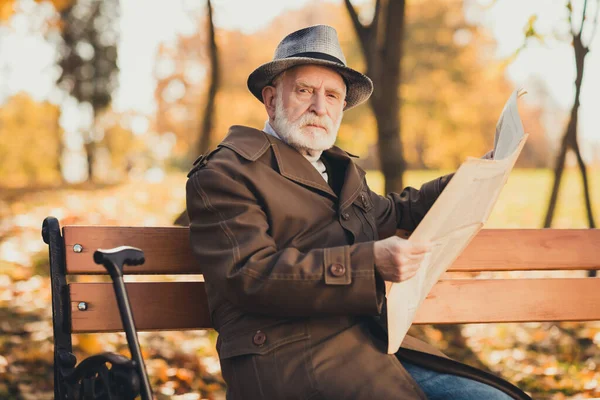 Foto av allvarlig gammal man grinig pensionär gillar inte barn spela höst stadspark sitta bänk läsa utseende tidning bära mössa lång jacka ytterkläder — Stockfoto