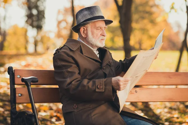 Foto del lado del perfil de un hombre viejo serio caballero inglés tienen parque mañana tiempo libre otoño caminar sentarse banco leer periódico usar gorra abrigo largo — Foto de Stock