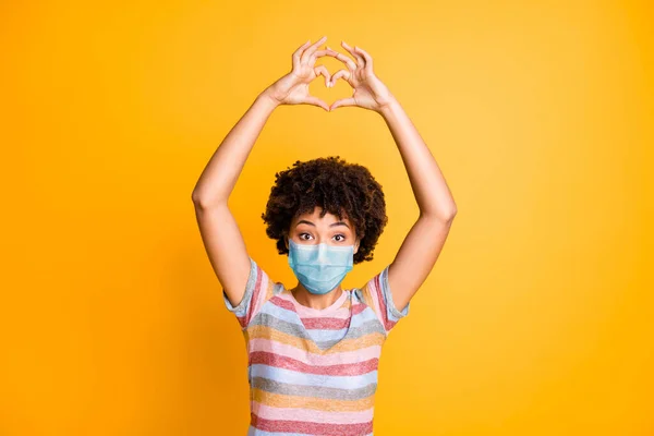 Retrato dela ela agradável menina de cabelos ondulados saudável vestindo máscara de gaze de segurança mostrando forma do coração medicina distância social isolado brilhante brilhante brilho vibrante cor amarela fundo — Fotografia de Stock